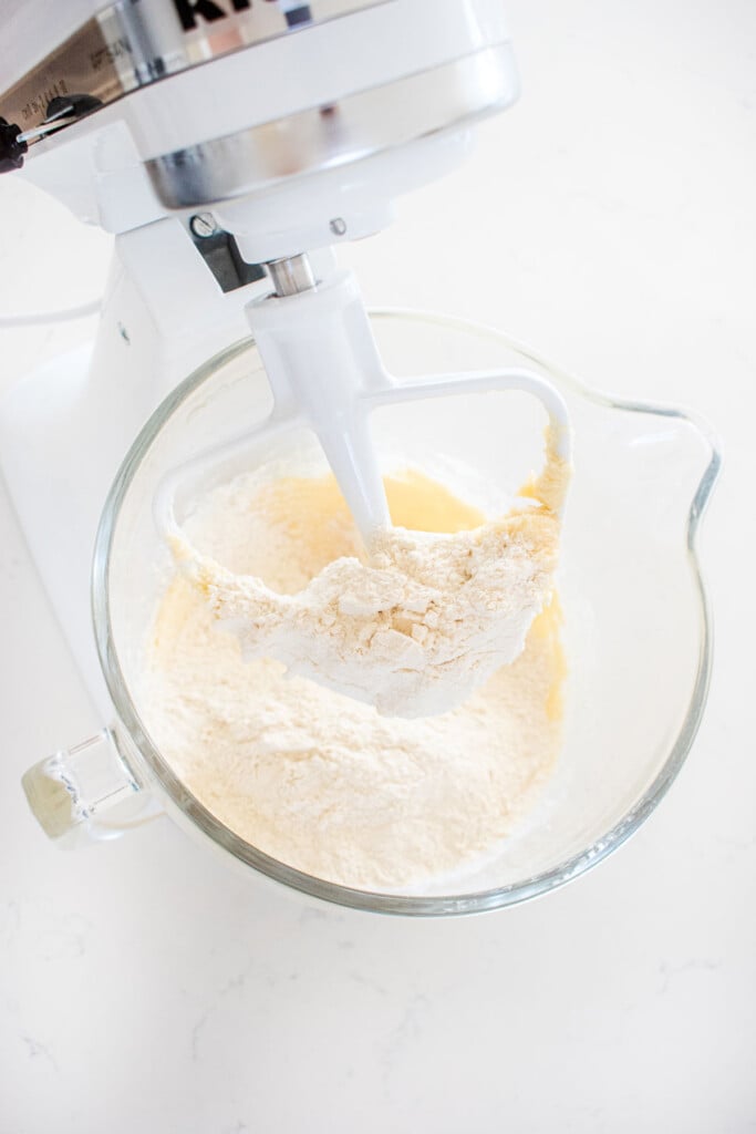 flour being added into a glass mixing bowl with easter egg thumbprint cookie dough on a marble counter.