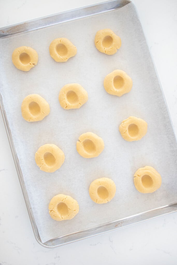 cookie dough balls with an indent pressed into the center on a baking sheet on a marble counter.