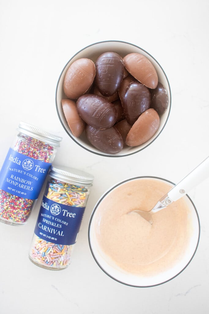 a bowl of chocolate easter eggs, a bowl of pink white chocolate, and sprinkle jars on a marble counter.