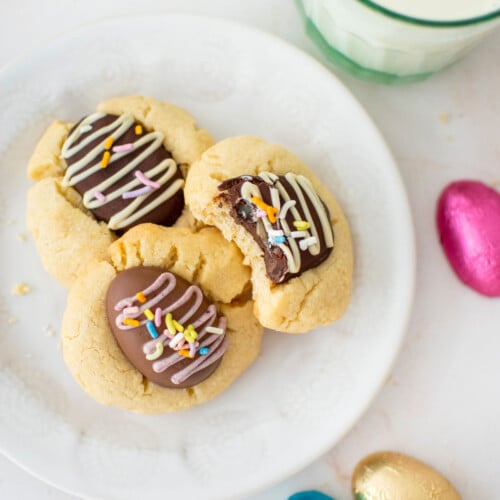 easter egg thumbprint cookies on a plate, one with a bite taken out with a glass of milk and chocolate eggs on a white marble counter.