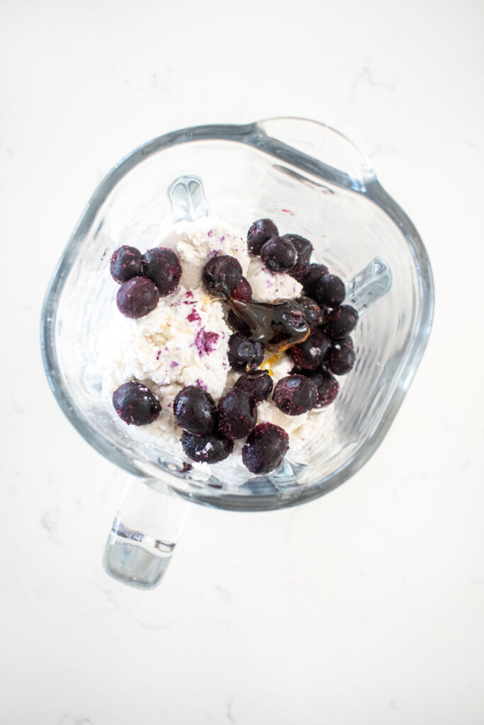 glass blender with coconut cream, frozen blueberries and honey on a white marble counter.