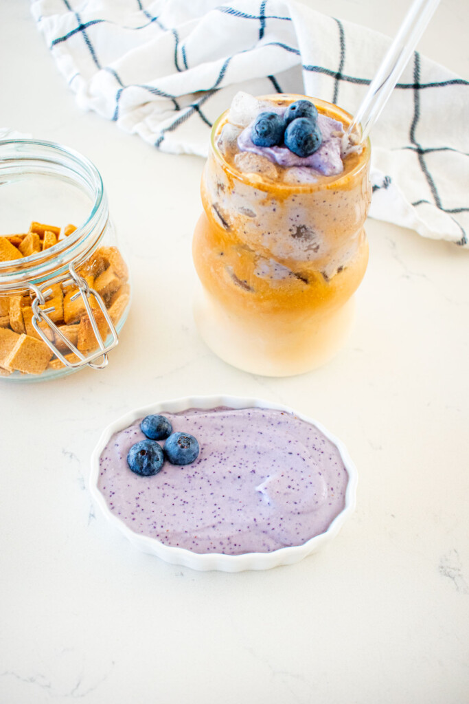 iced latte with blueberry cold foam, blueberry pup cup and dog treats on a white marble counter.