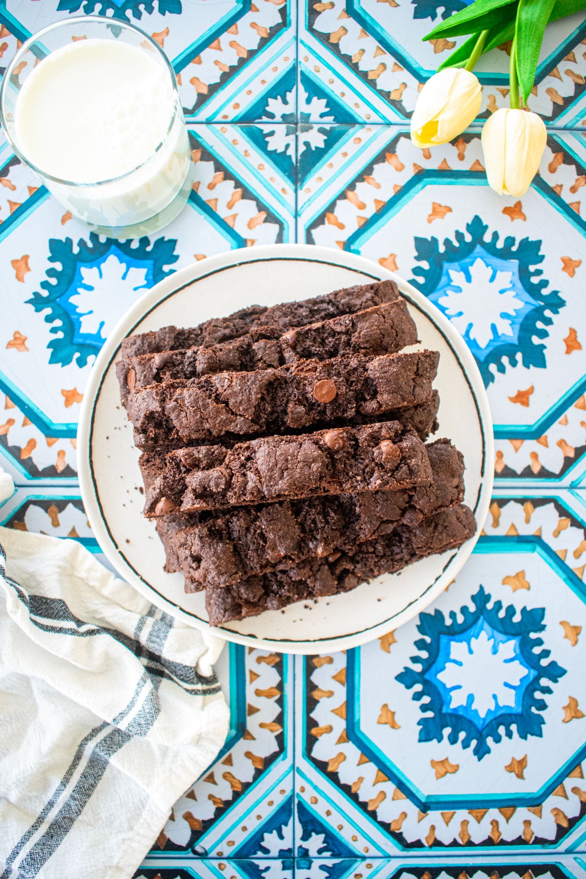 simple chocolate espresso biscotti piled onto a plate on a tile table with a glass of milk and fresh tulips.