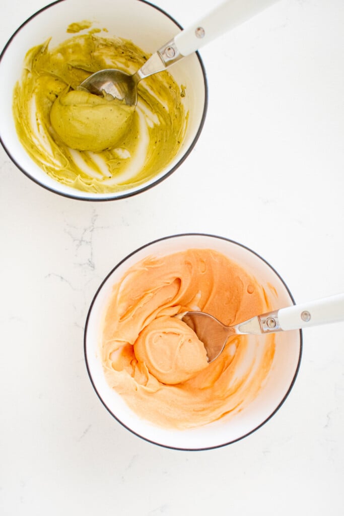 two small bowls with light orange frosting and light green frosting on a white marble counter.