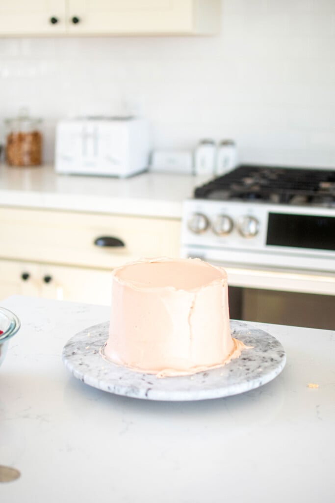 light pink strawberry frosted on a layer cake on a marble cake turntable on a white marble counter.