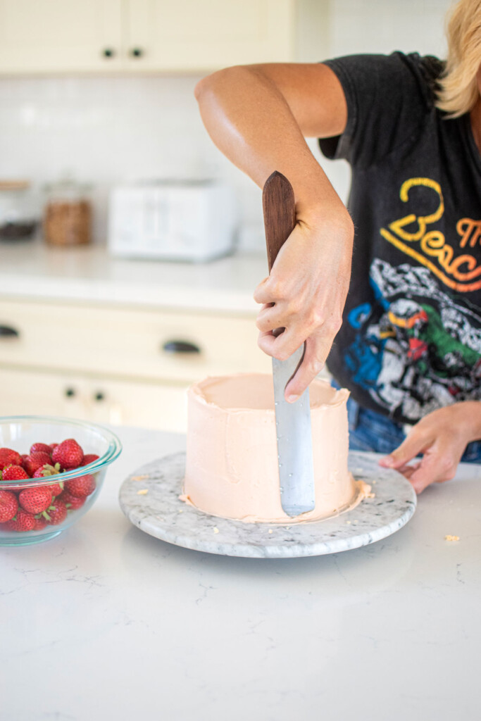 woman frosting a strawberry cake with light pink frosting on a marble turnbable at the kitchen counter.