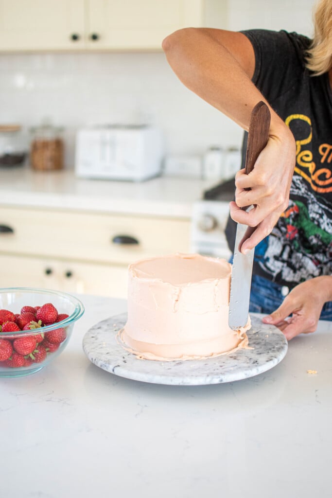woman spiraling a strawberry layer cake on a cake turntable on a marble counter in the kitchen.