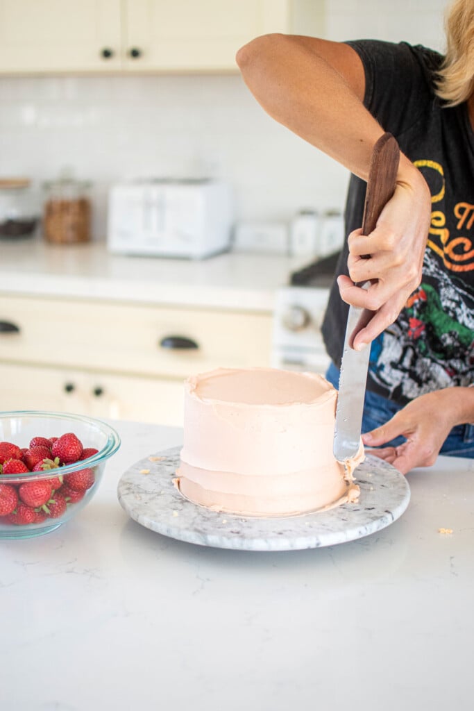 woman spiraling a light pink buttercream cake on a cake turntable on a white marble counter.