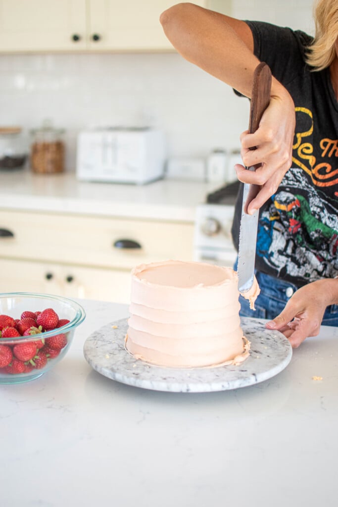 woman spiraling a light pink buttercream cake on a cake turntable on a white marble counter.