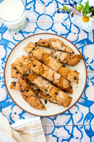 blueberry muffin biscotti on a plate on a blue tile table with fresh flowers and a glass of milk.