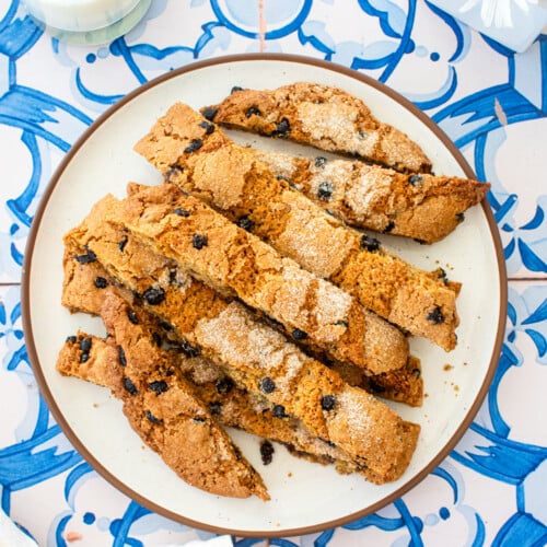 blueberry muffin biscotti on a plate on a blue tile table with fresh flowers and a glass of milk.