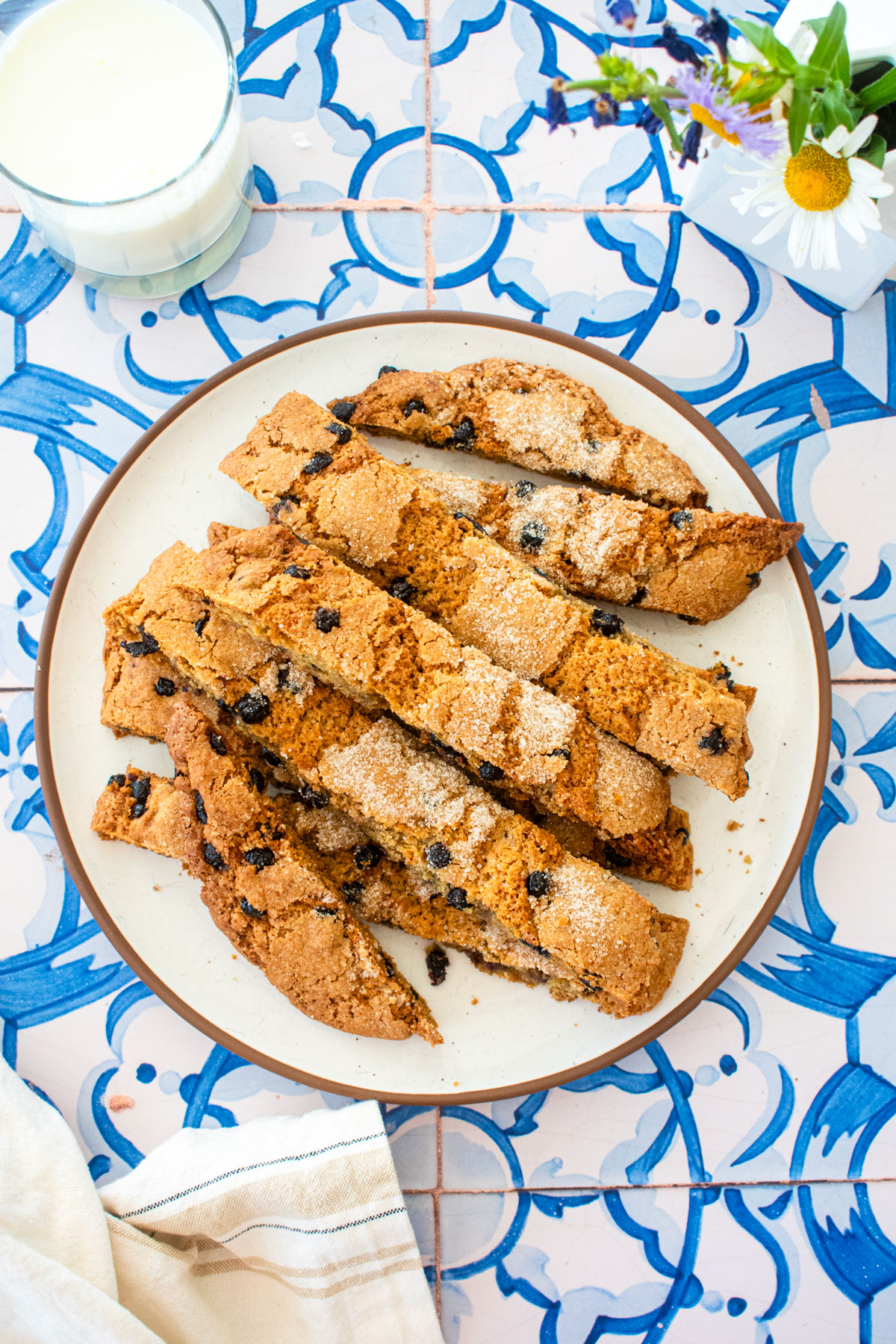 blueberry muffin biscotti on a plate on a blue tile table with fresh flowers and a glass of milk.