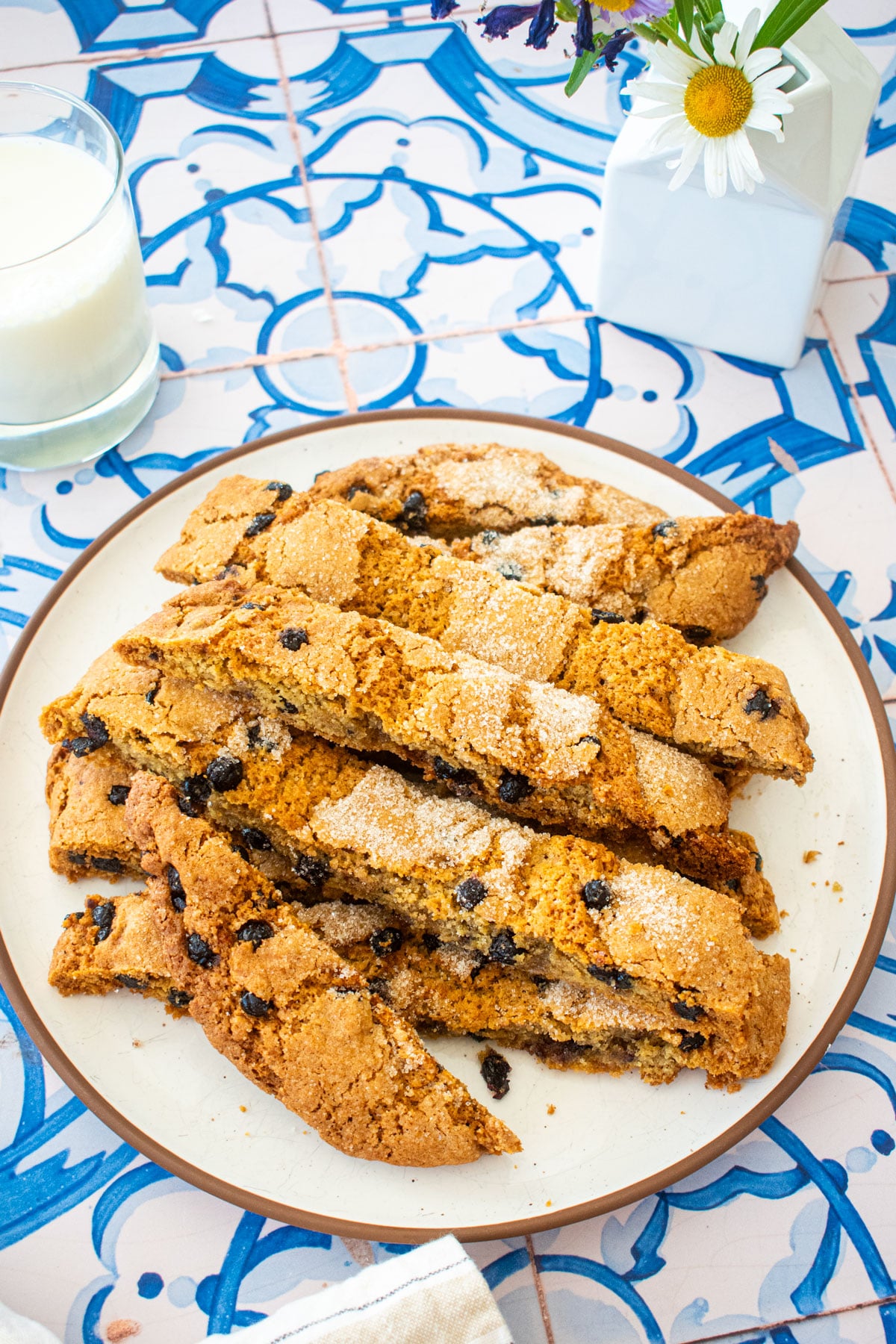 blueberry muffin biscotti on a plate on a blue tile table with fresh flowers and a glass of milk.