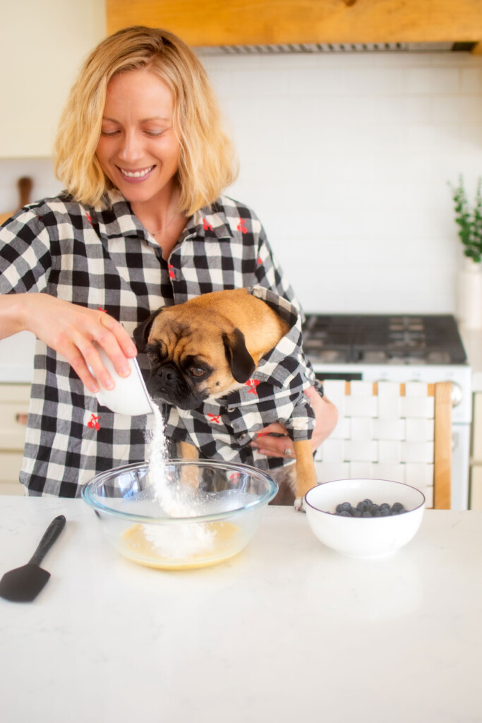 woman and pug in kitchen making blueberry pancakes for dogs, pouring flour into mixing bowl.