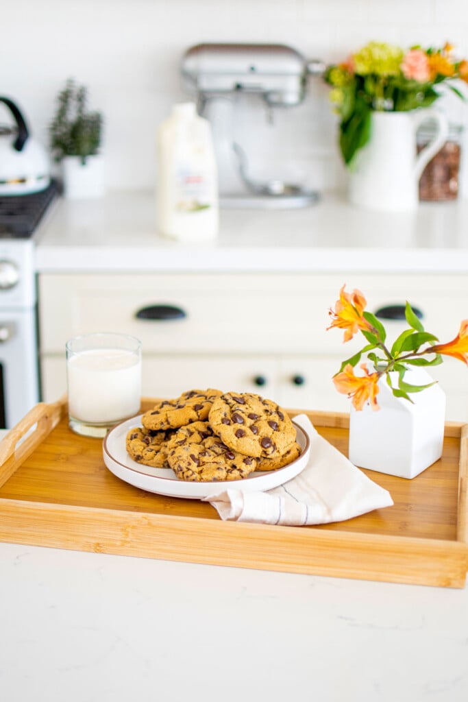 wood tray with a plate of cream cheese chocolate chip cookies and a glass of milk and fresh flowers on a white kitchen counter.