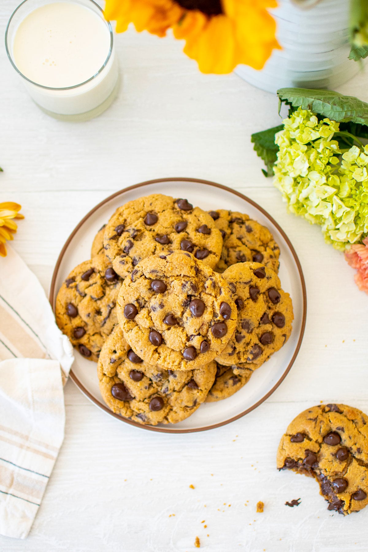 cream cheese chocolate chip cookies on a plate on a white wood table with a glass of milk and fresh flowers.