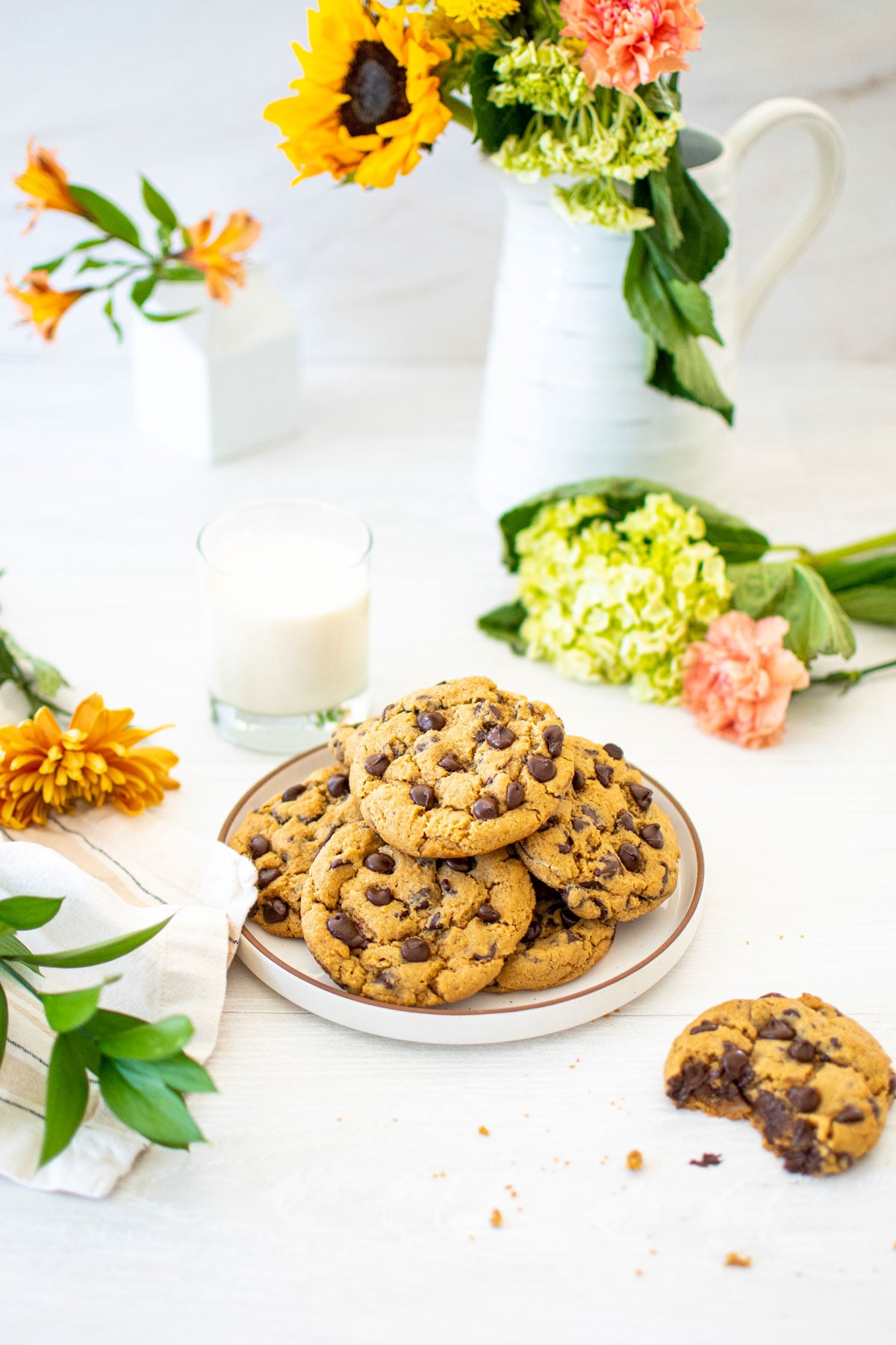 plate of cream cheese chocolate chip cookies on a white wood table with fresh flowers and a glass of milk.