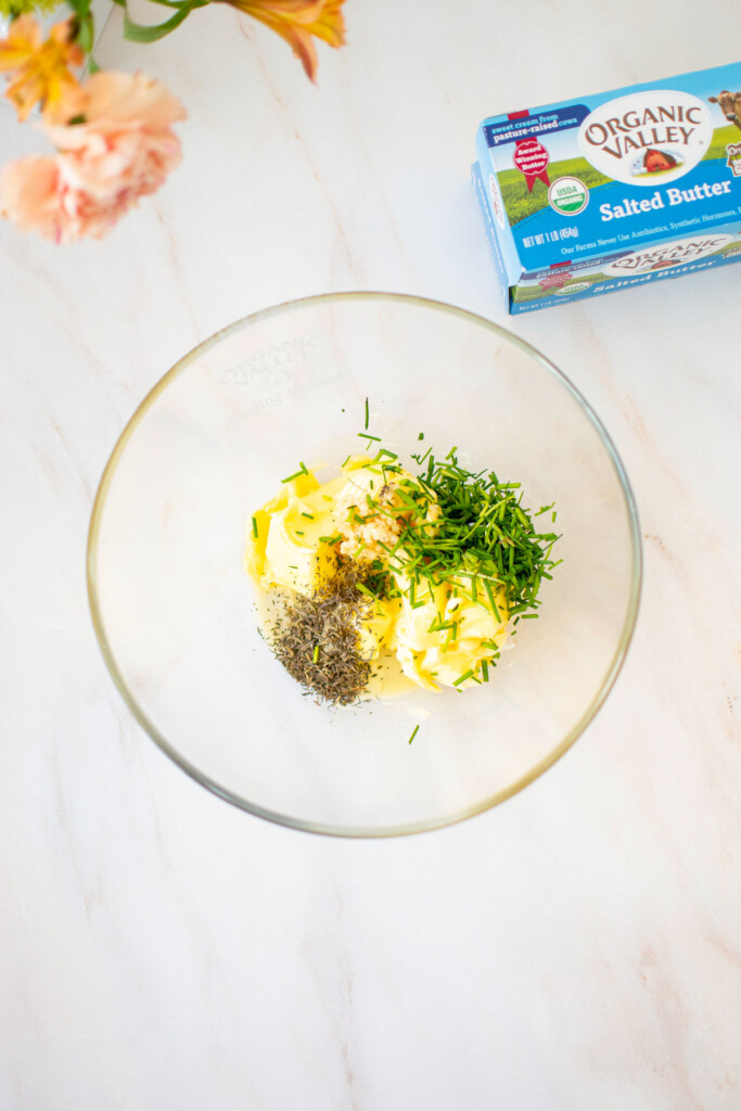 ingredients for garlic herb butter in a glass mixing bowl on a marble counter.
