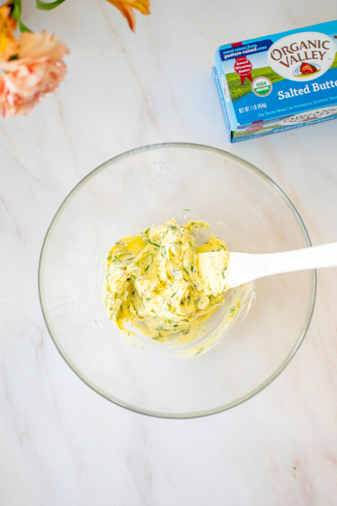 garlic herb butter in a glass mixing bowl with a white spatula on a marble counter.