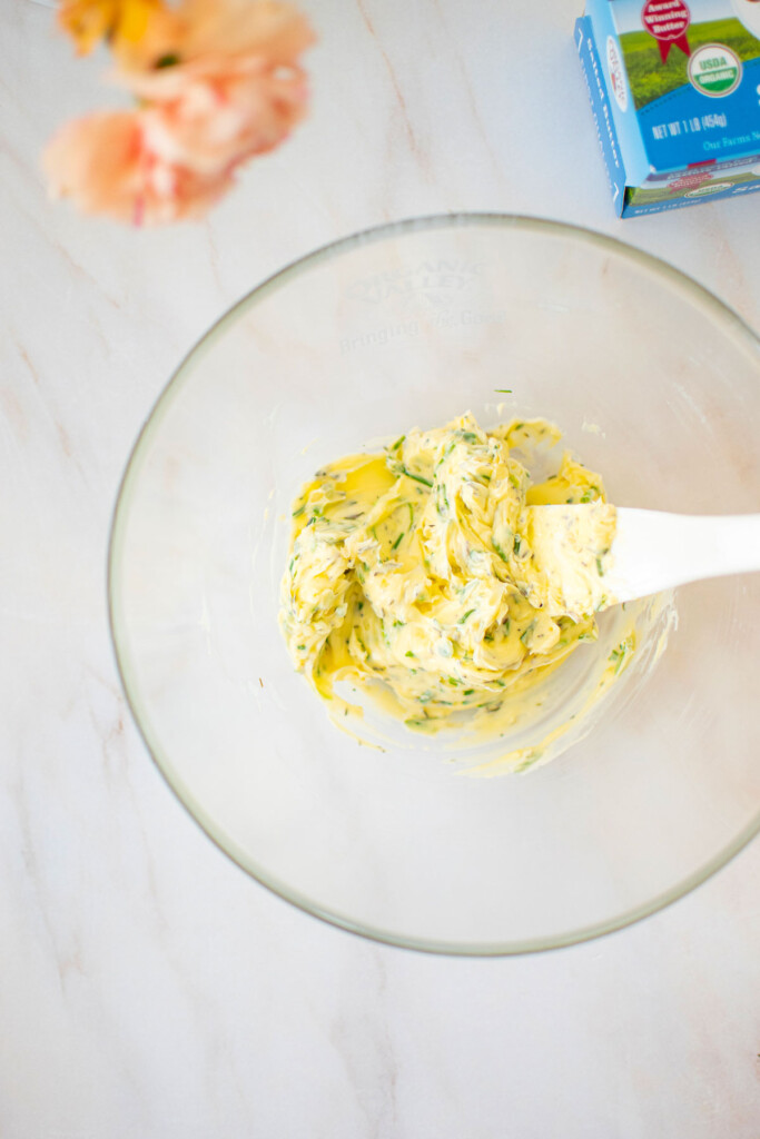 garlic herb butter in a glass mixing bowl with a white spatula on a marble counter.