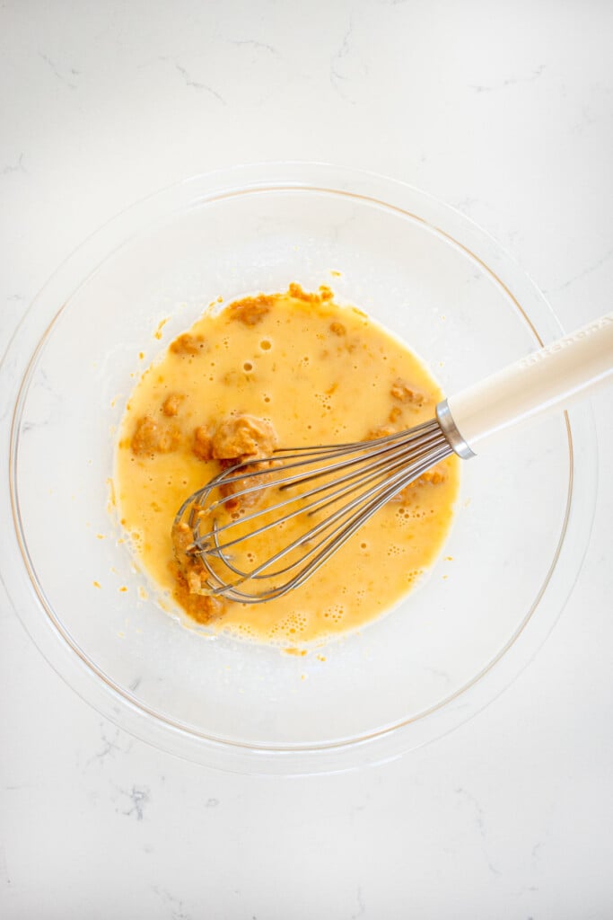 wet ingredients for blueberry peanut butter dog cake in a glass mixing bowl with a whisk on a white marble countertop.
