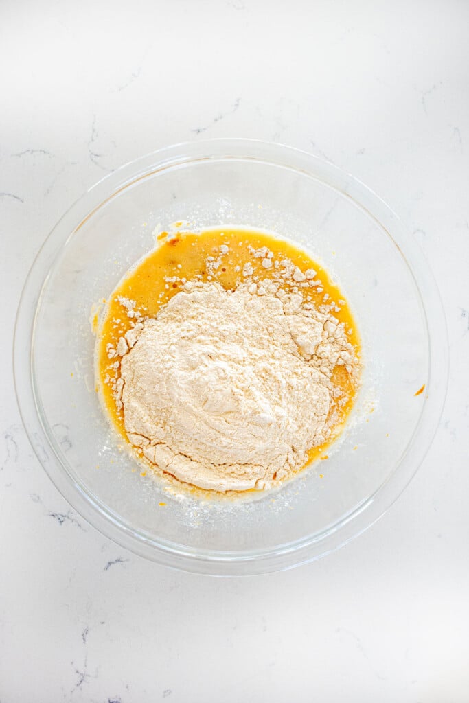 wet and dry ingredients for blueberry peanut butter dog cake in a glass mixing bowl on a white marble countertop.