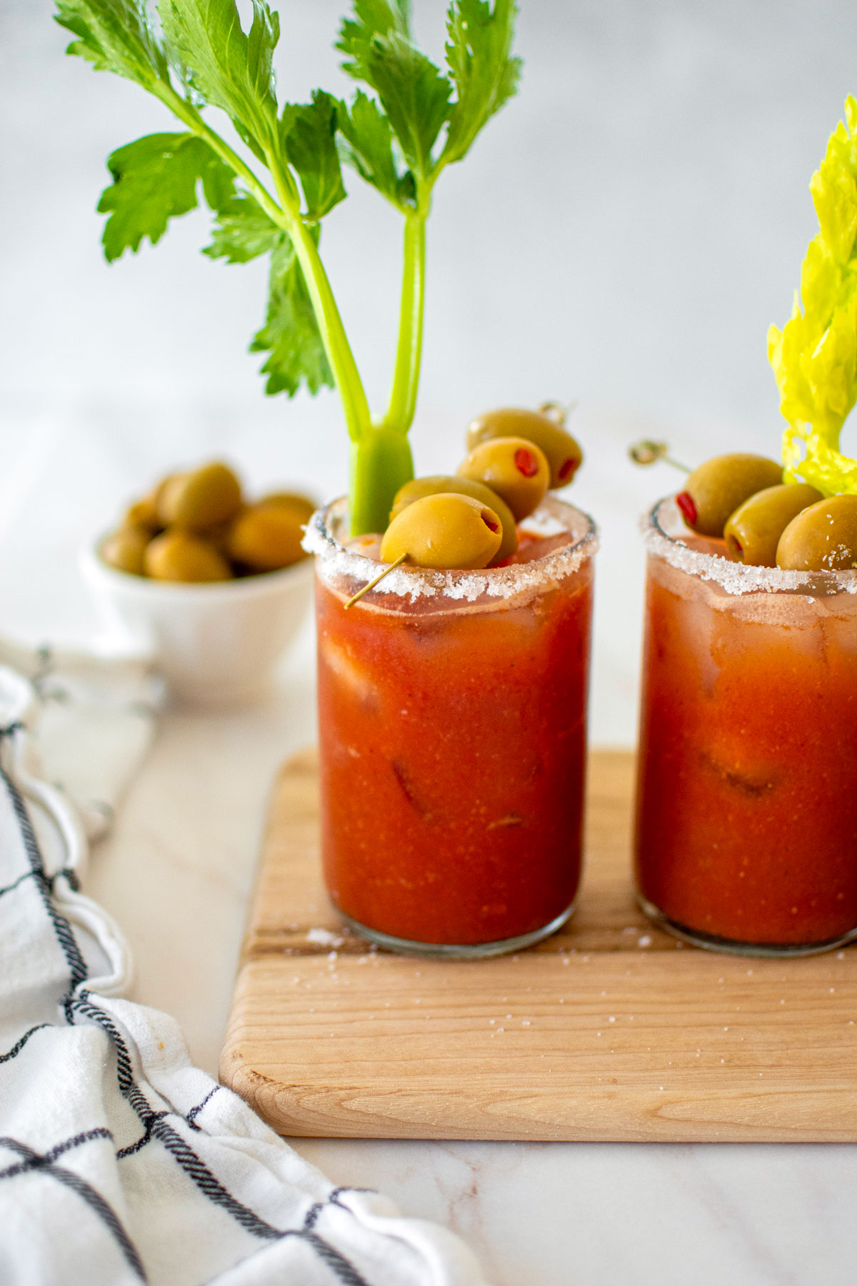 bone broth bloody marys with green olives and celery on a white marble counter.