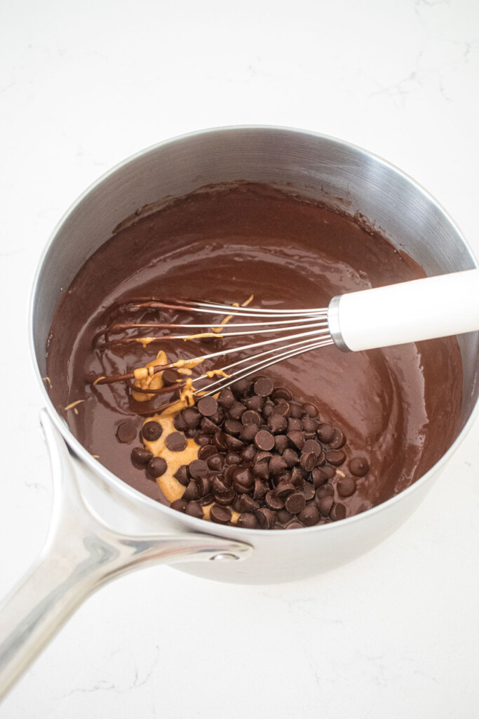 eggless chocolate peanut butter pudding with chocolate chips and peanut butter being added to it with a whisk on a marble counter.