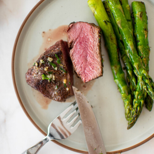 medium rare garlic herb beef tenderloin sliced in half on a plate with asparagus on a marble countertop.