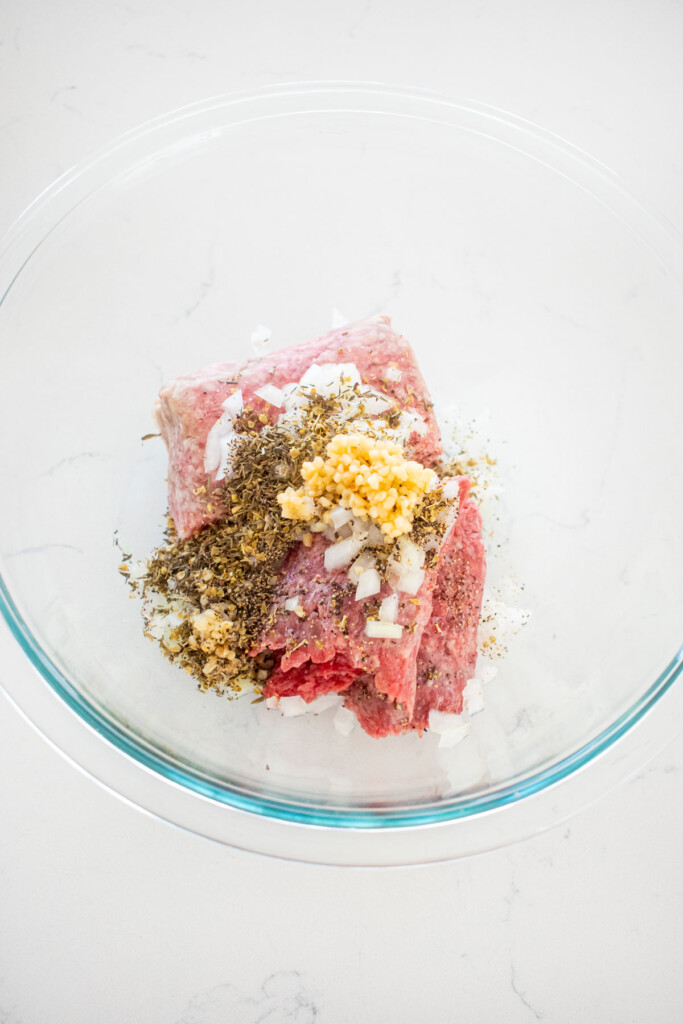glass bowl of ground beef and lamb with spices on a white marble counter.