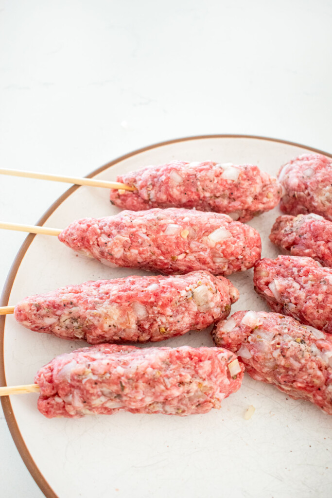 raw ground beef and lamb mixture formed around skewers on a plate on a marble counter.
