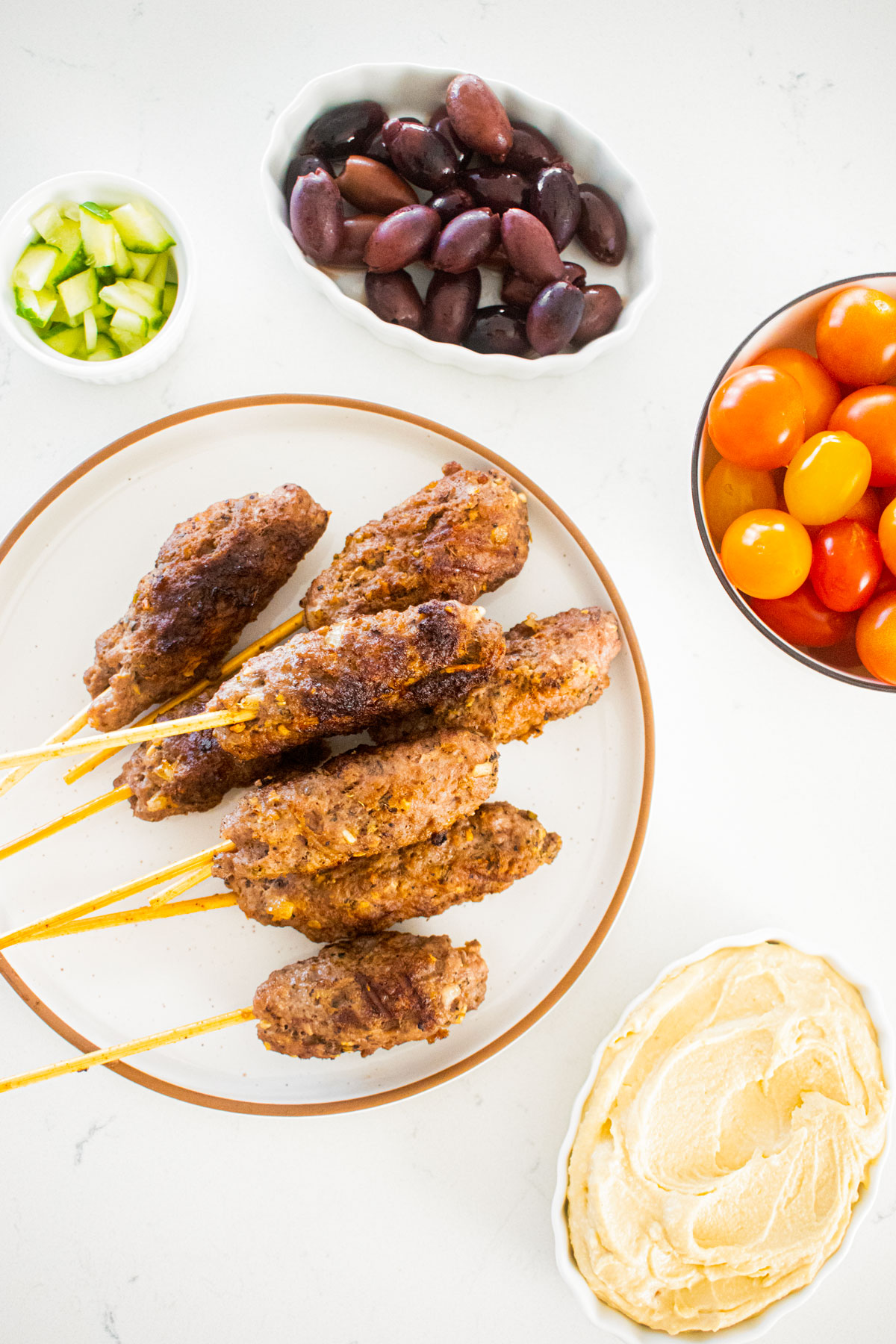 beef and lamb kabobs on a plate with cherry tomatoes, kalamata olives, cucumber and hummus dishes next to it on a marble counter.