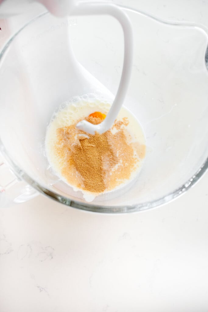 glass stand mixing bowl with milk, egg, yeast, sourdough discard, and cane sugar on a white marble counter.