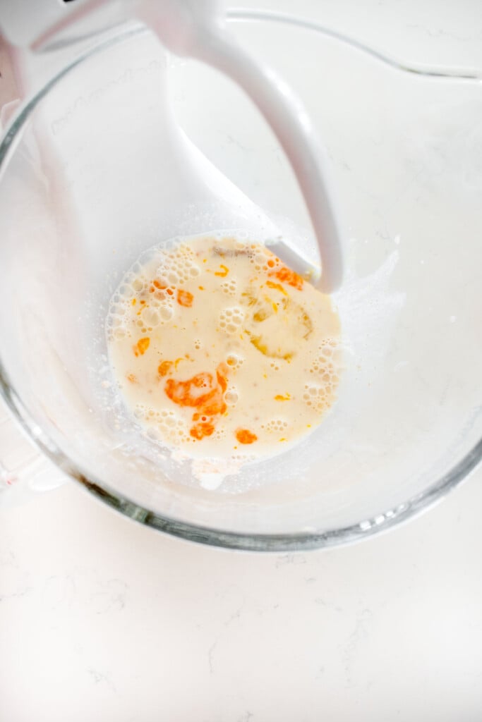 glass stand mixing bowl with mixed milk, egg, yeast, sourdough discard, and cane sugar on a white marble counter.