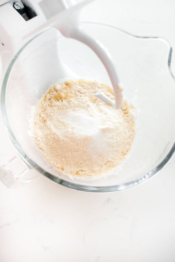 dry ingredients for homemade sourdough hot dog buns added into wet ingredients in a glass stand mixing bowl on a white marble counter.