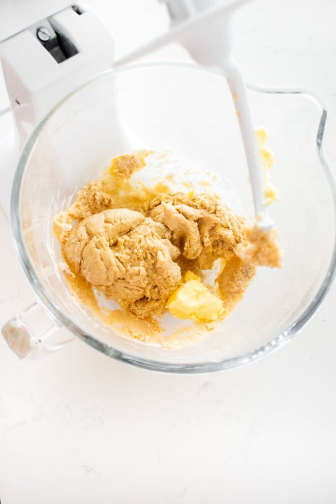 mixed sourdough hot dog bun dough with butter in a glass stand mixing bowl on a white marble counter.