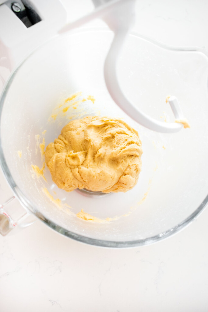 mixed sourdough hot dog bun dough in a glass stand mixing bowl on a white marble counter.