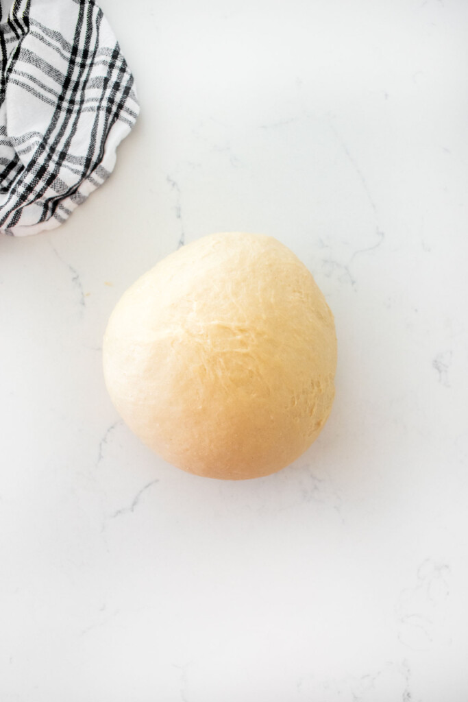 homemade sourdough hot dog bun dough rolled into a smooth ball on a white marble counter.