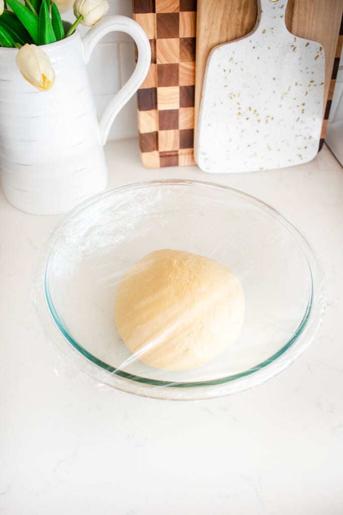 homemade sourdough hot dog bun dough in a glass mixing bowl covered with plastic wrap on a white marble counter.