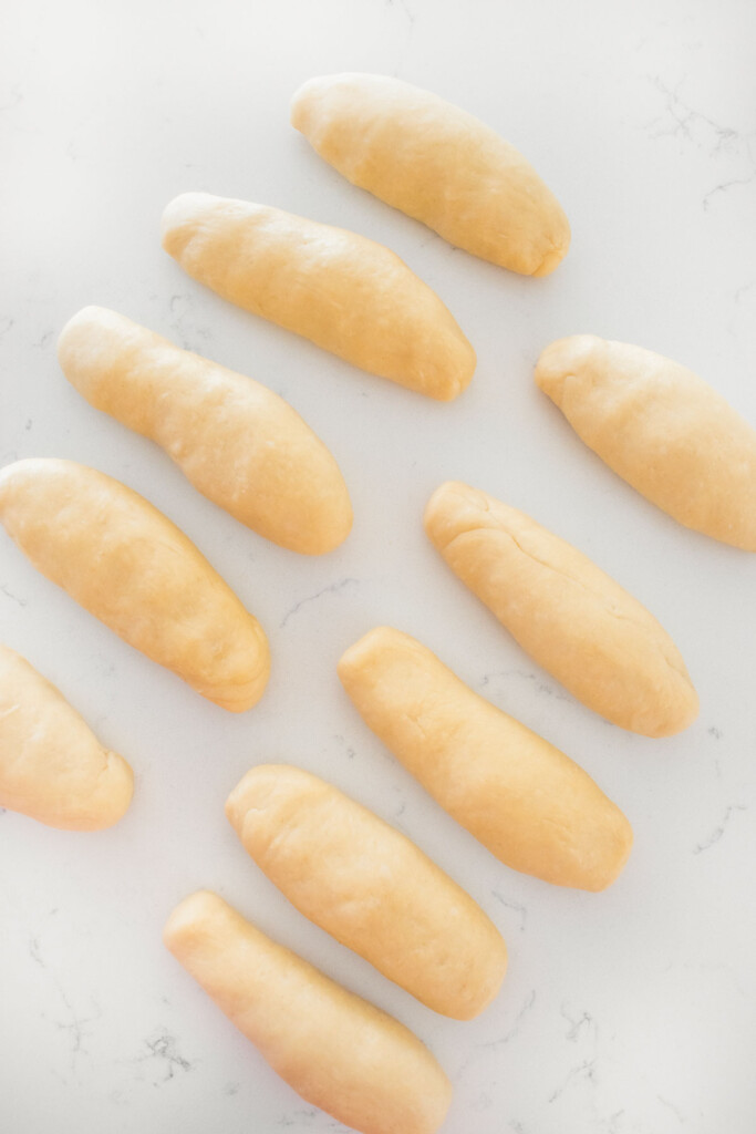 homemade sourdough hot dog bun dough rolled into small ropes on a white marble counter.