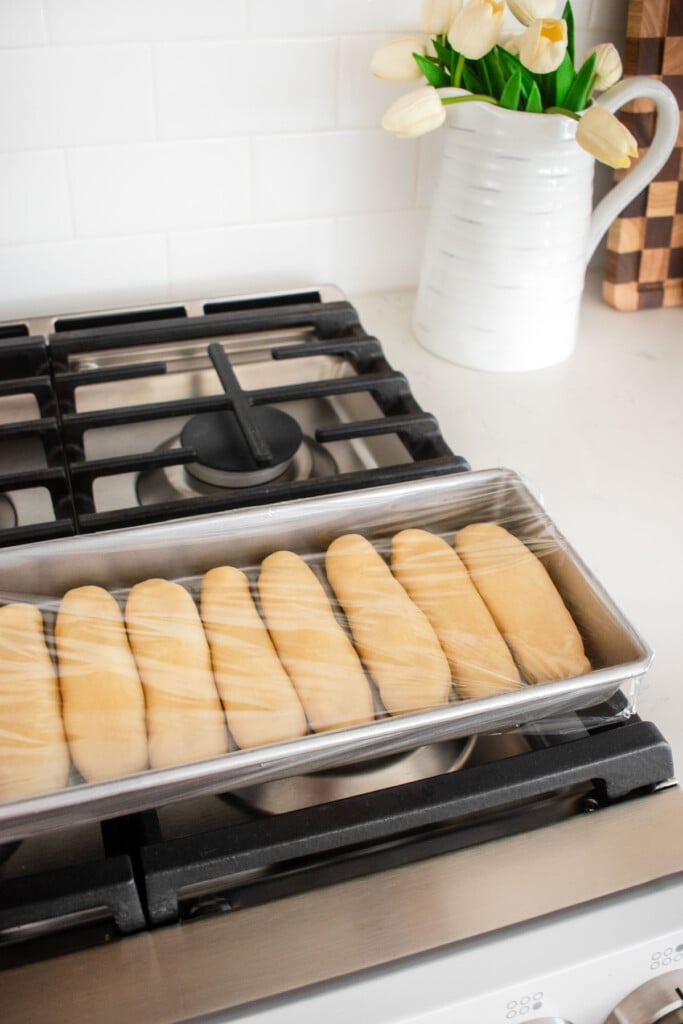 homemade sourdough hot dog bun dough in a hot dog pan covered with plastic wrap proofing on top of an oven.