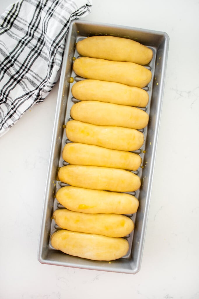 homemade sourdough hot dog bun dough in a hot dog pan brushed with egg on a white marble counter.