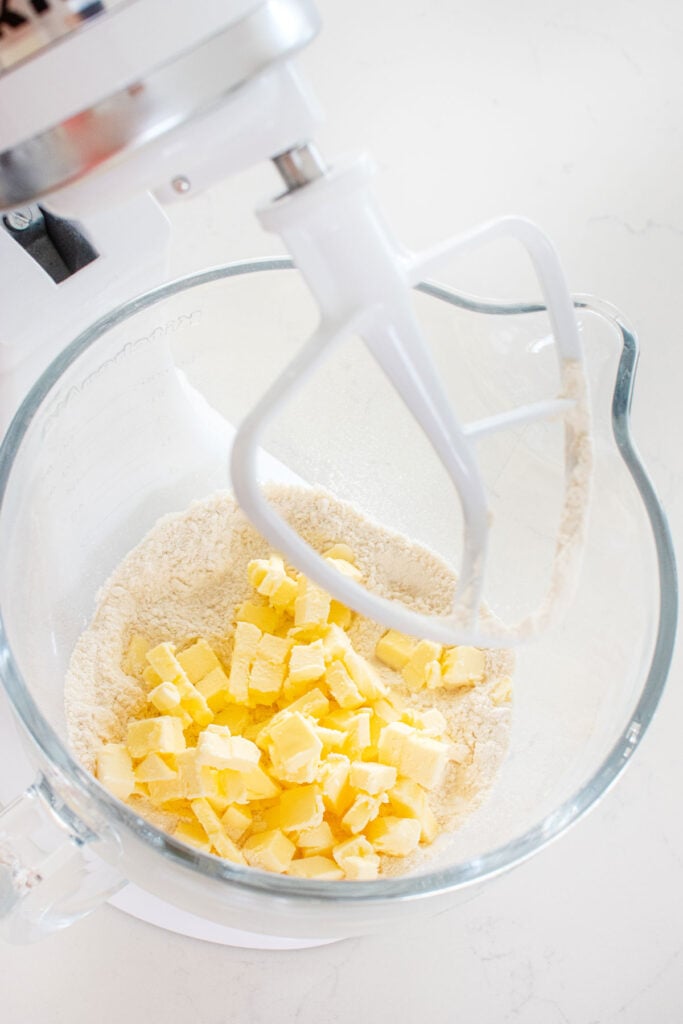 flour, sugar and chopped butter in a mixing bowl of a stand mixer on a white marble countertop.