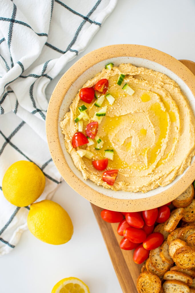 lemon tahini hummus in a bowl on a wood cutting board with veggies and bagel chips.