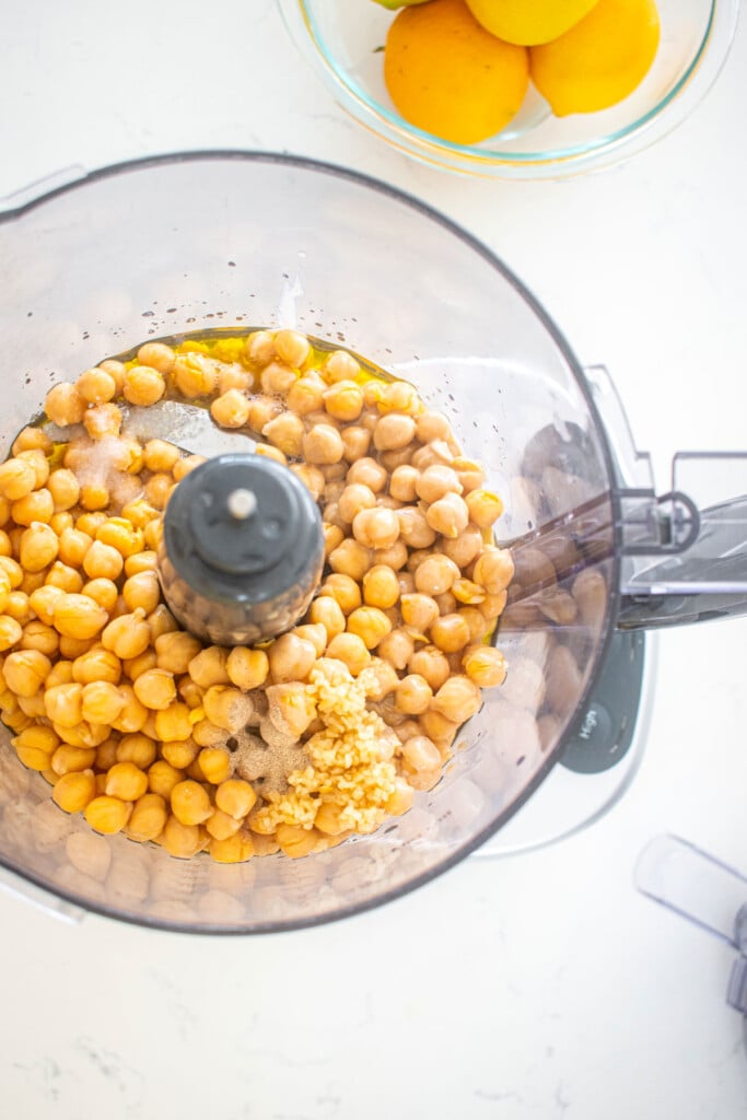 ingredients for lemon tahini hummus in a food processor on a white marble countertop.