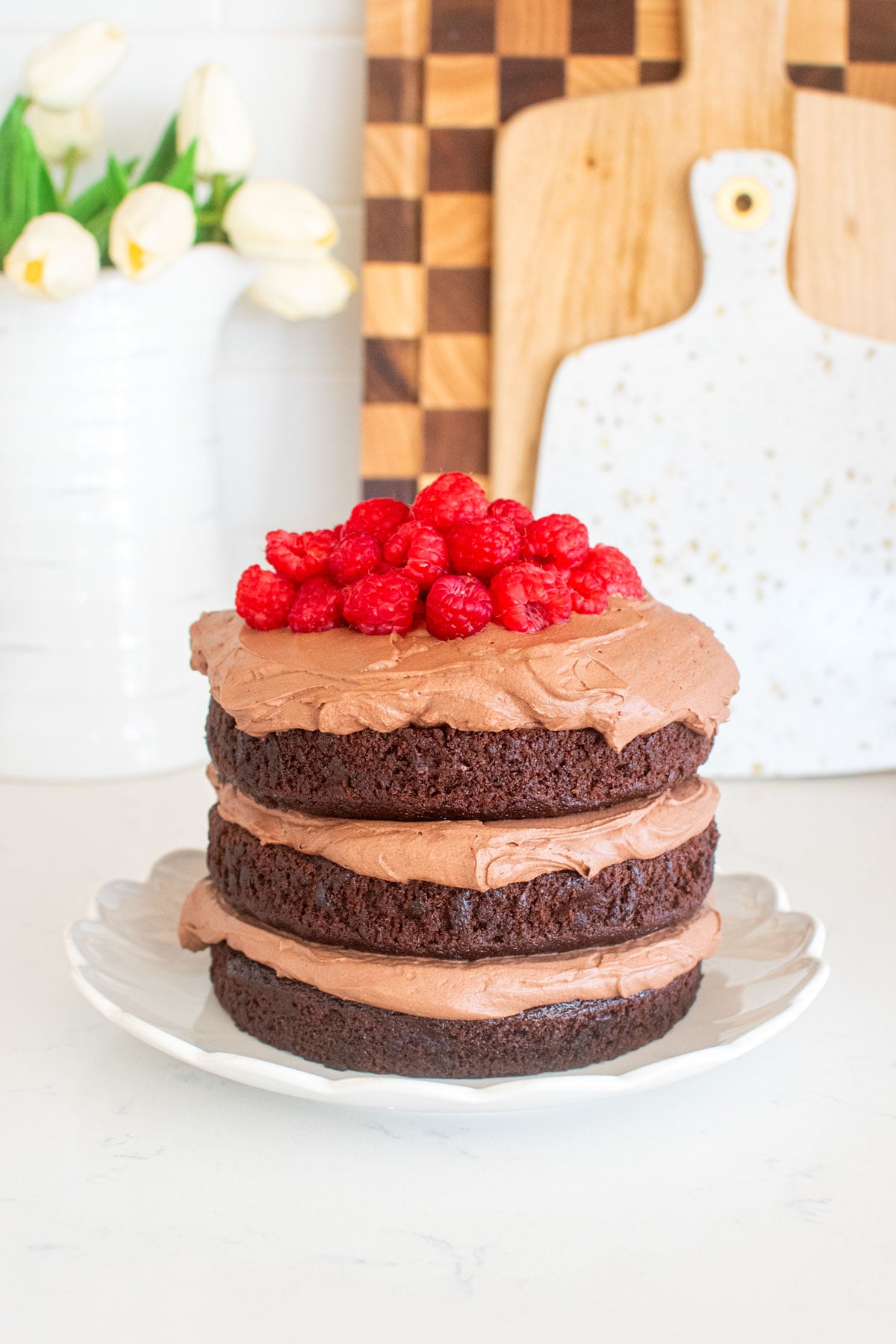 chocolate naked cake with chocolate whipped cream frosting and fresh raspberries on top on a white marble counter.