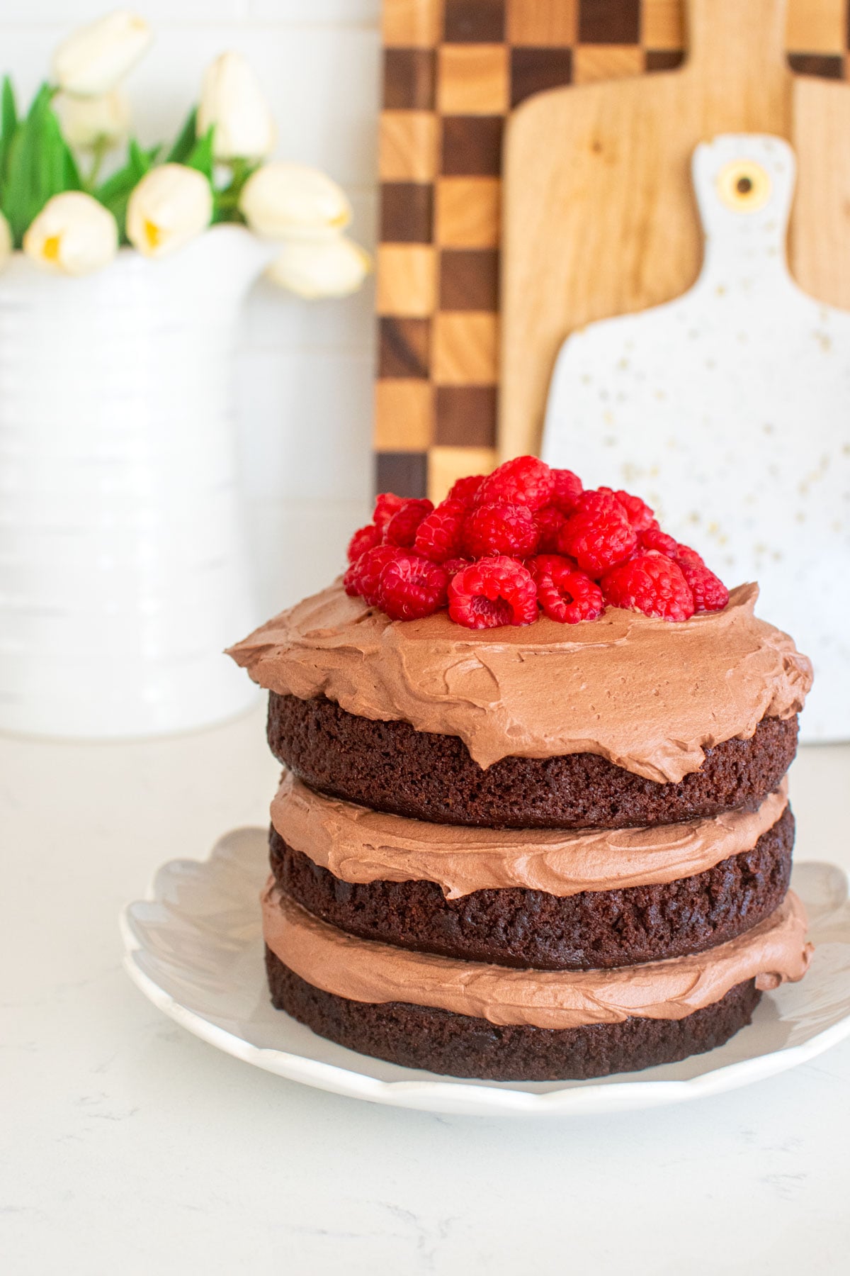 chocolate naked cake with chocolate whipped cream frosting and fresh raspberries on top on a white marble counter.