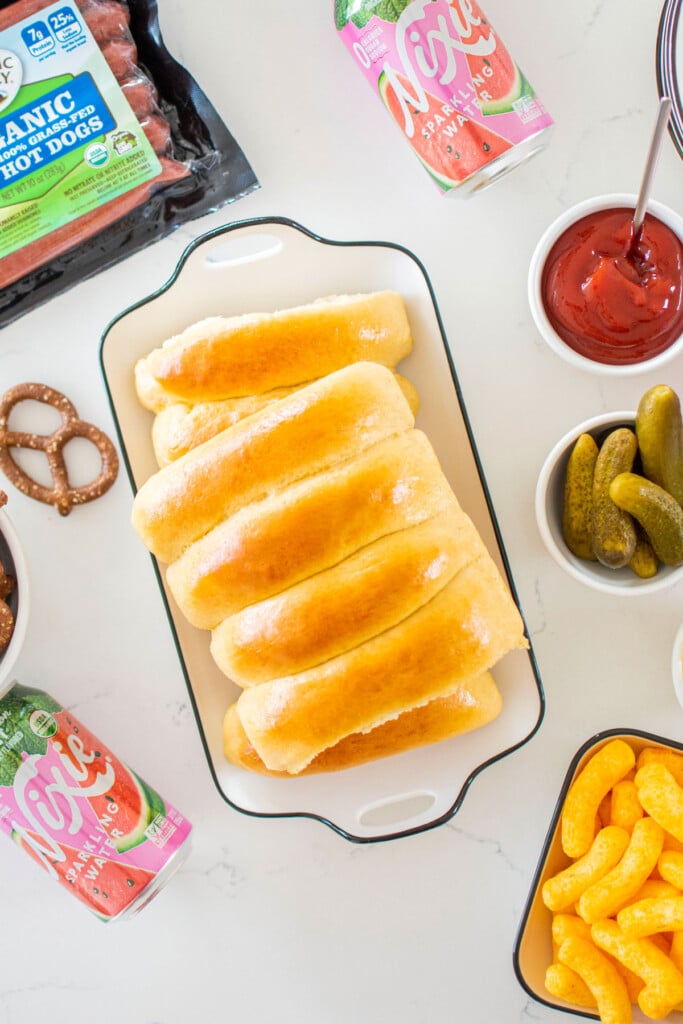 homemade sourdough hot dog buns on a platter with hot dog toppings around.