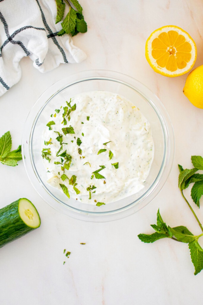 tzatziki sauce with mint on top in a glass bowl on a marble countertop with lemons and cucumber.