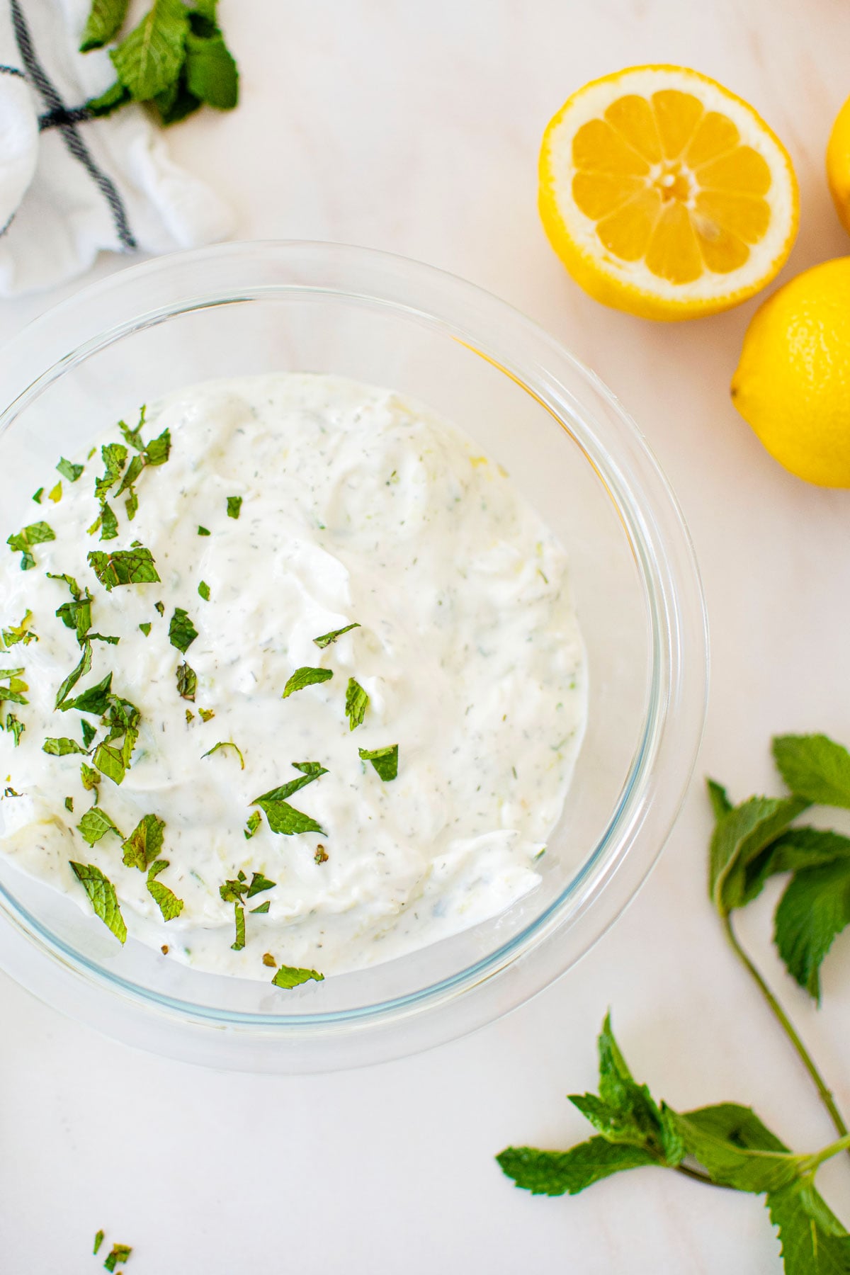 tzatziki sauce with mint on top in a glass bowl on a marble countertop with lemons and cucumber.